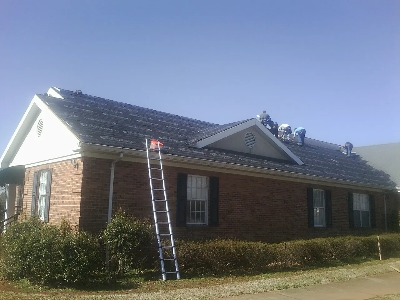 Crew tearing shingles off a brick ranch roof with a ladder set — Emergency Roof Repair work by our crew in Gillett