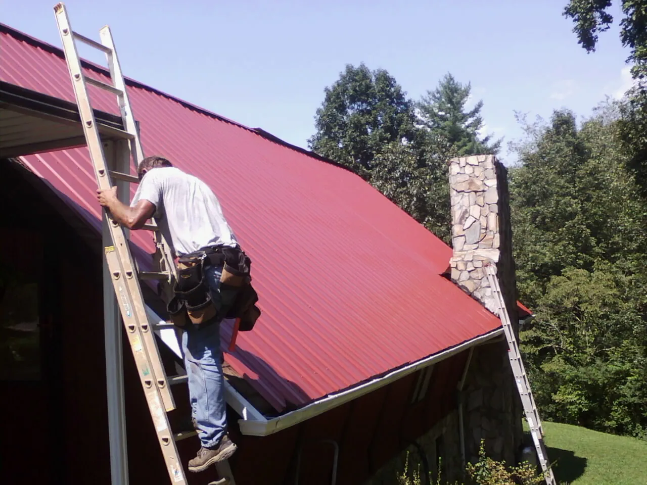 Roofer on a ladder beside a red metal roof and stone chimney — Soffit & Fascia Repair by Roofing Brothers in Gillett